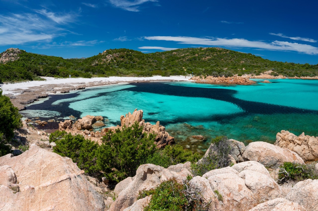 Luftaufnahme einer Meeresbucht mit türkisblauem, klarem Wasser. Der Strand ist von feinem Sand bedeckt und von grüner Vegetation sowie Felsen umgeben. 