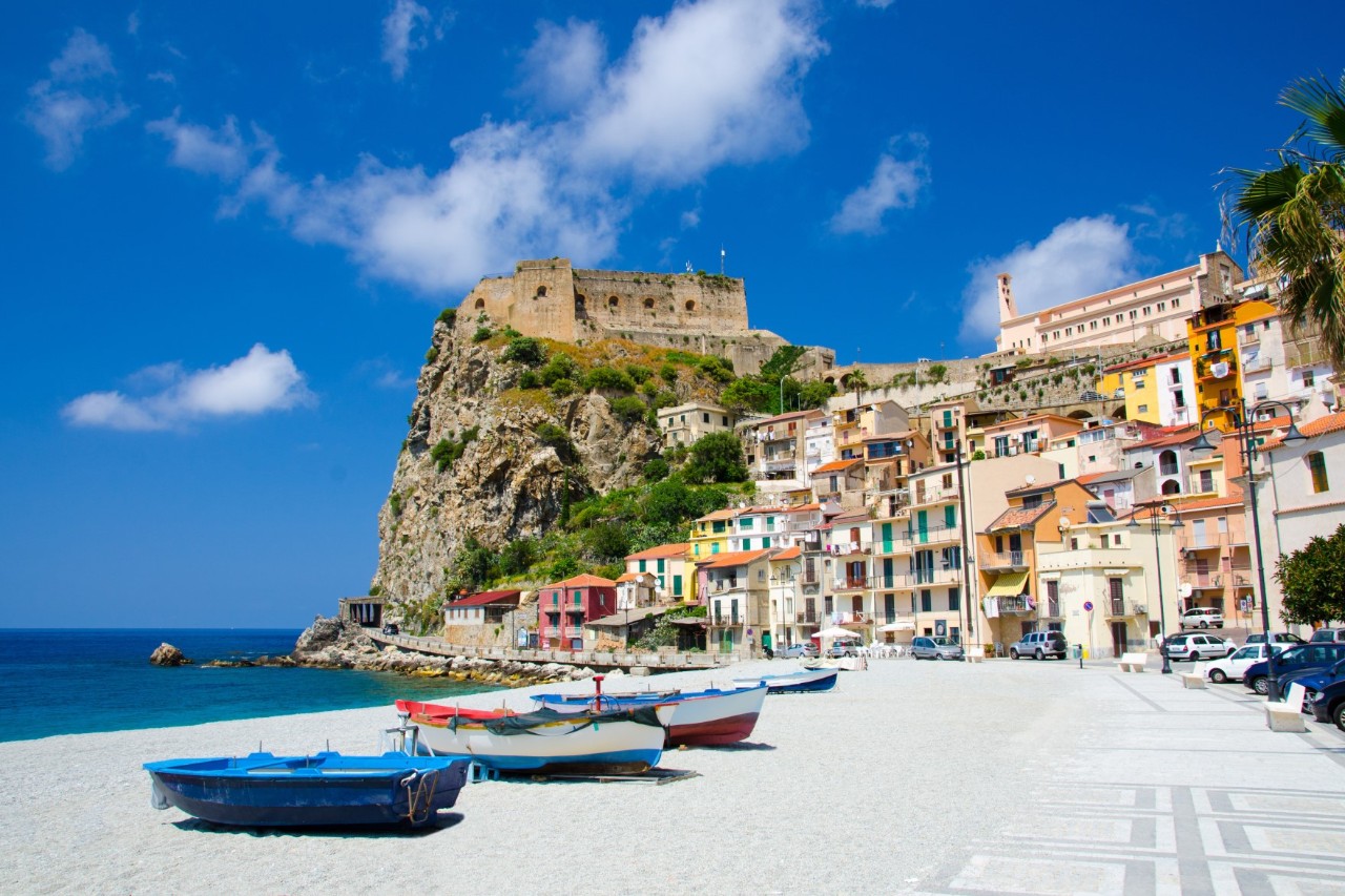 Mediterrane Küstenstadt mit bunten Häusern, die an einen Felsen gebaut sind. Auf dem Felsen thront eine historische Festung. Im Vordergrund ist ein schmaler Strandabschnitt mit Fischerbooten im Sand, dahinter das blaue Meer. 