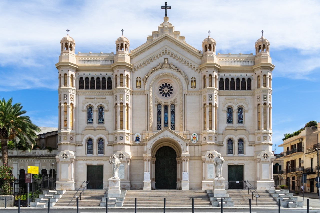Vorderansicht einer Kathedrale mit heller Fassade, gestaltet im neoromanischen und neugotischen Stil. Sie verfügt über vier symmetrische Türme, eine große Rosette oberhalb des Eingangs und detaillierte Verzierungen, flankiert von Statuen, die auf der Treppe stehen.