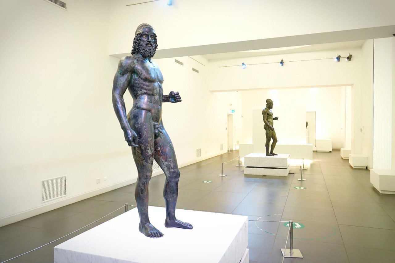 Männliche, muskulöse Bronzestatue im Vordergrund. Eine weitere Statue steht im Hintergrund. Diese sind auf Podesten in einem hellen Museumsraum ausgestellt. 