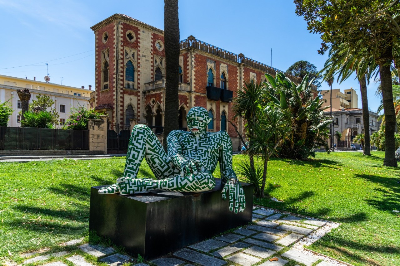 Skulptur eines menschlichen Körpers mit einem labyrinthartigen Muster, die auf einem schwarzen Sockel sitzt. Im Hintergrund steht ein neugotisch gestaltetes Gebäude mit rot-weißer Fassade. Palmen und Grünflächen umgeben die Szene.