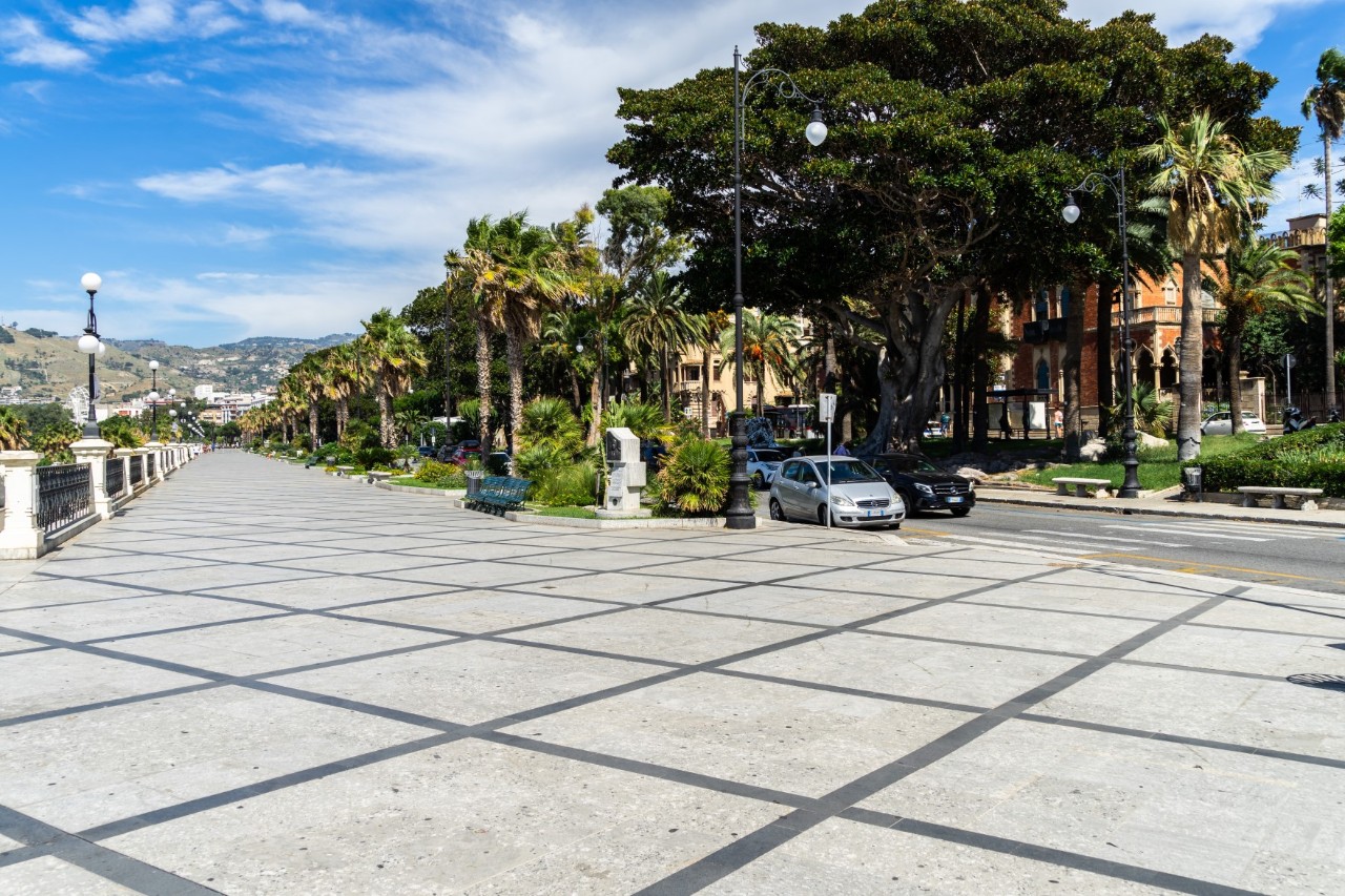 Beiter, gepflasterter Weg mit Schachbrettmuster, der von Palmen und Laternen gesäumt ist. Auf der rechten Seite befindet sich eine Straße, parkende Autos und ein kleiner Park mit großen Bäumen.