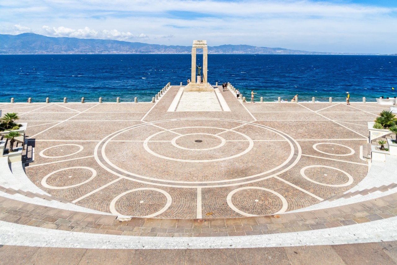 Großer Platz, der mit geometrischen Mustern verziert ist. Am Ende des Platzes steht ein Torbogen mit einer Statue. Dahinter erstreckt sich das tiefblaue Meer, eingerahmt von einer bergigen Küste im Hintergrund.