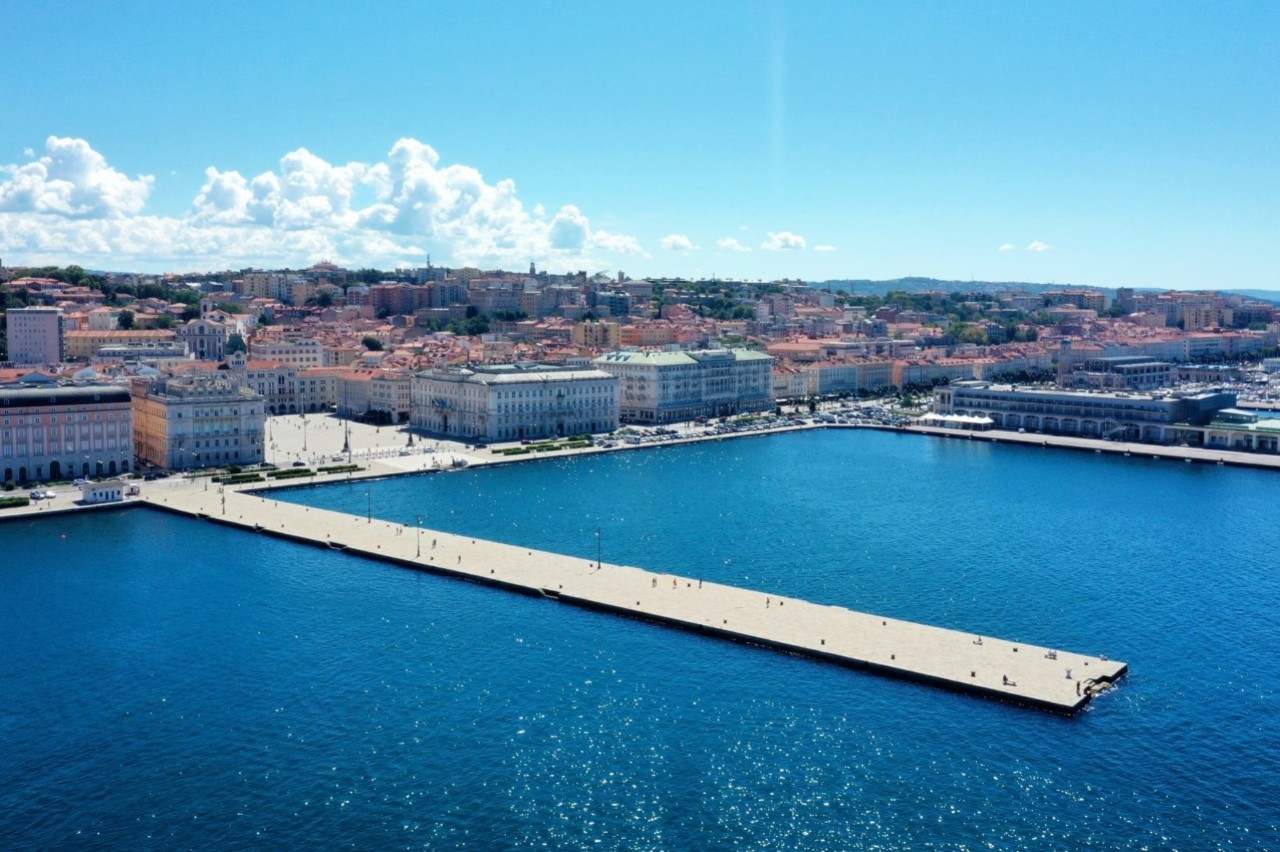 Luftaufnahme des ins Meer verlaufenden Kais mit Stadtpromenade, dahinter Stadtansicht, leicht bewölkter Himmel.
