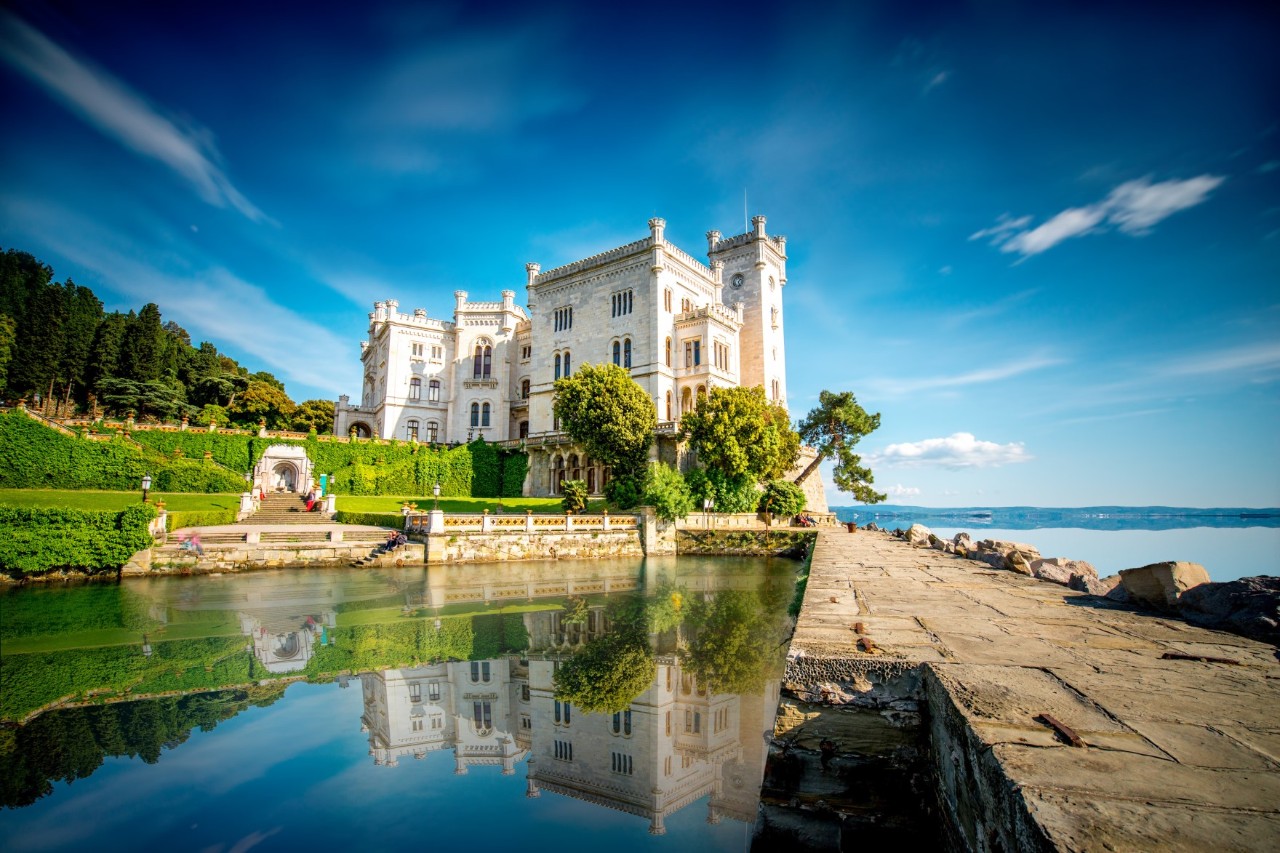 Weißes Schloss am Meer mit Grünanlage und Steinpromenade. Es spiegelt sich in einem Wasserbecken.