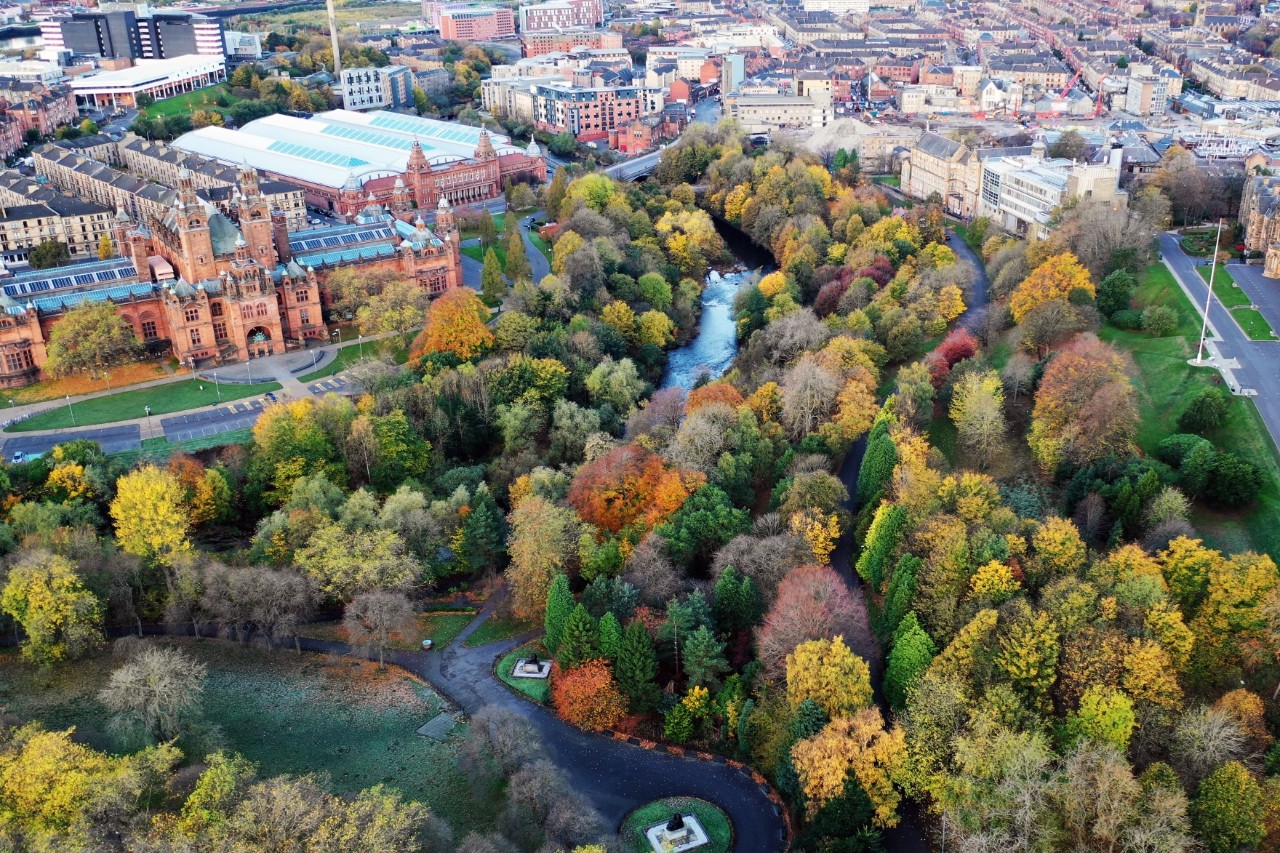 Eine urbane Landschaft aus der Vogelperspektive. Im Vordergrund ist ein großer Park mit einem Fluss und Bäumen in herbstlichen Farben zu sehen. Rechts im Bild sind Häuser zu sehen. Links im Bild befindet sich ein markantes Gebäude im viktorianischen Stil mit roten Backsteinen.