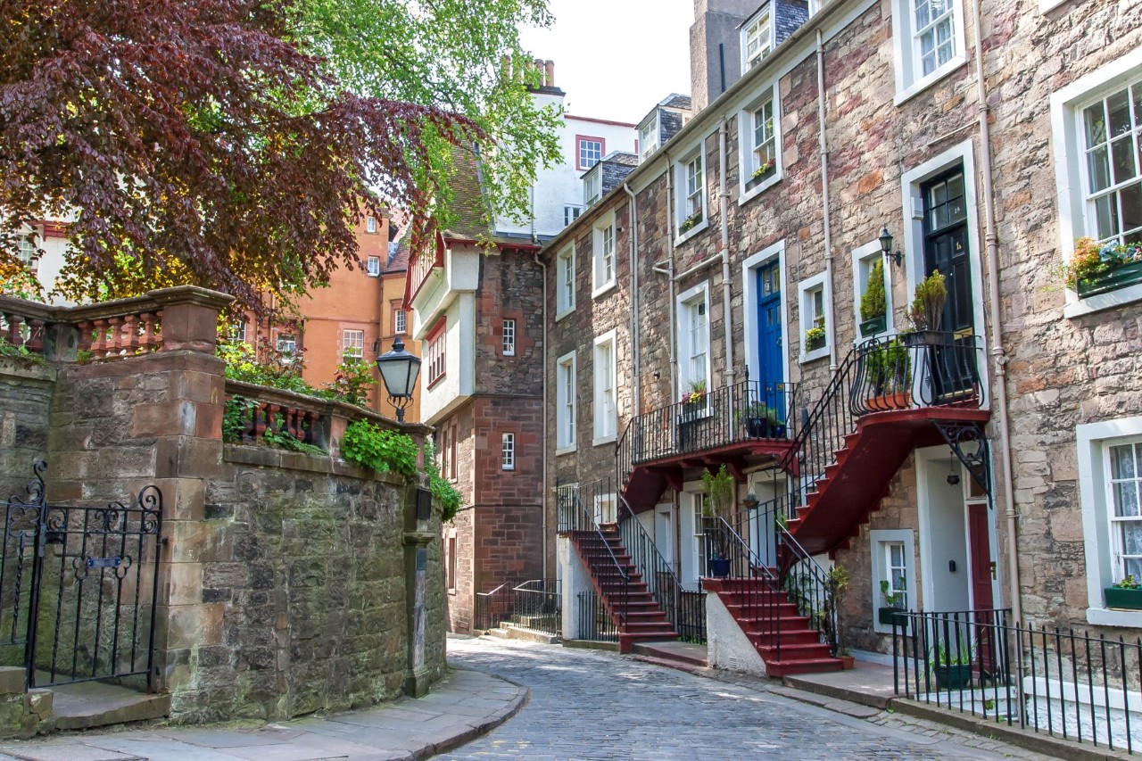 Eine ruhige Gasse mit Reihenhäusern aus Stein. Die Häuser haben bunte Türen, schmiedeeiserne Balkone und Außentreppen. Links ist eine alte Steinmauer mit einem schmiedeeisernen Tor zu sehen, umgeben von üppiger Vegetation.