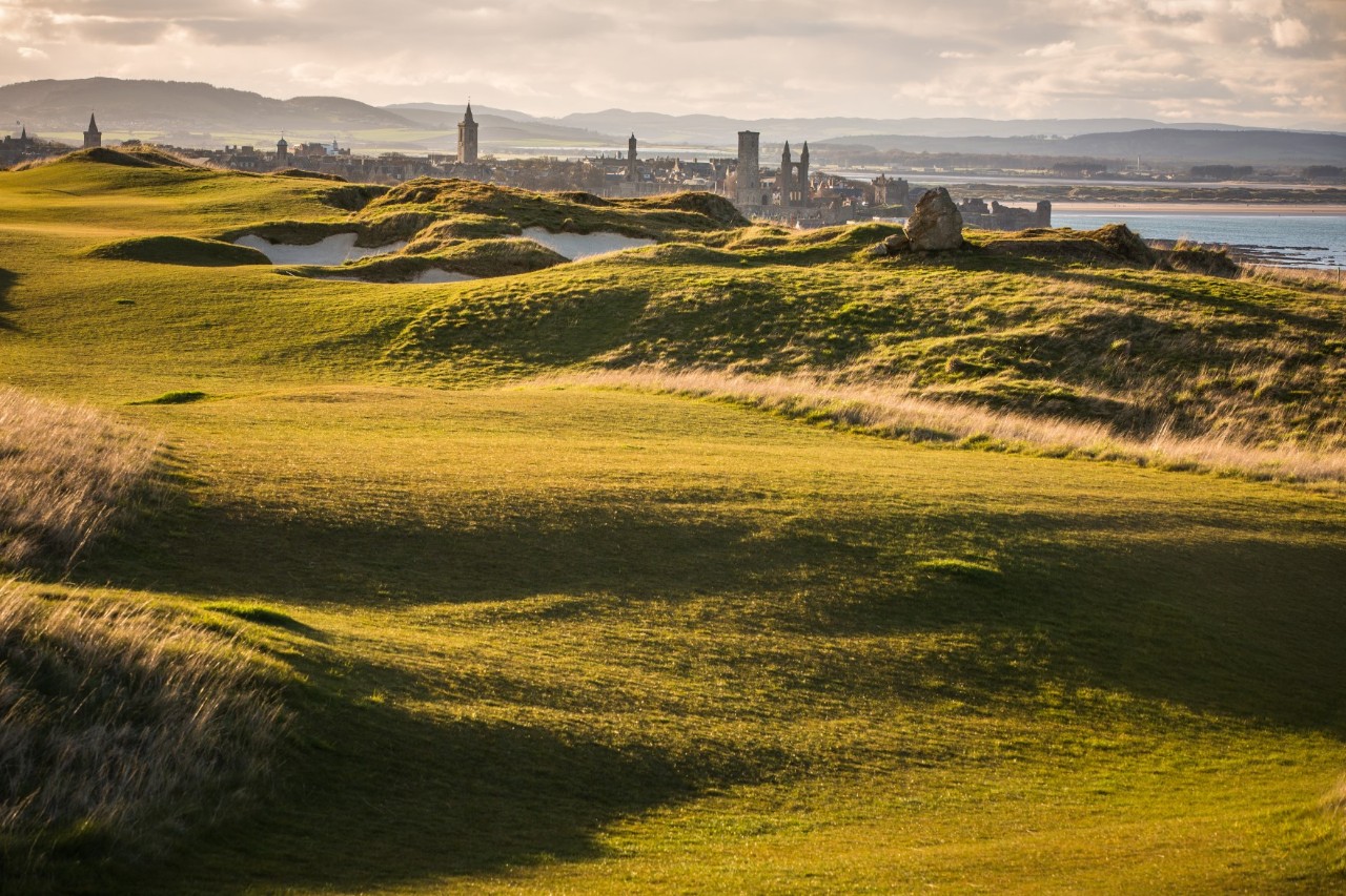 Eine hügelige, grasbewachsene Landschaft, die einem Golfplatz ähnelt. Klein im Hintergrund sind eine Küstenlinie und eine Stadt mit alten Türmen und historischen Gebäuden zu sehen. Der Himmel ist wolkig und dunstig in warmen Rosatönen.