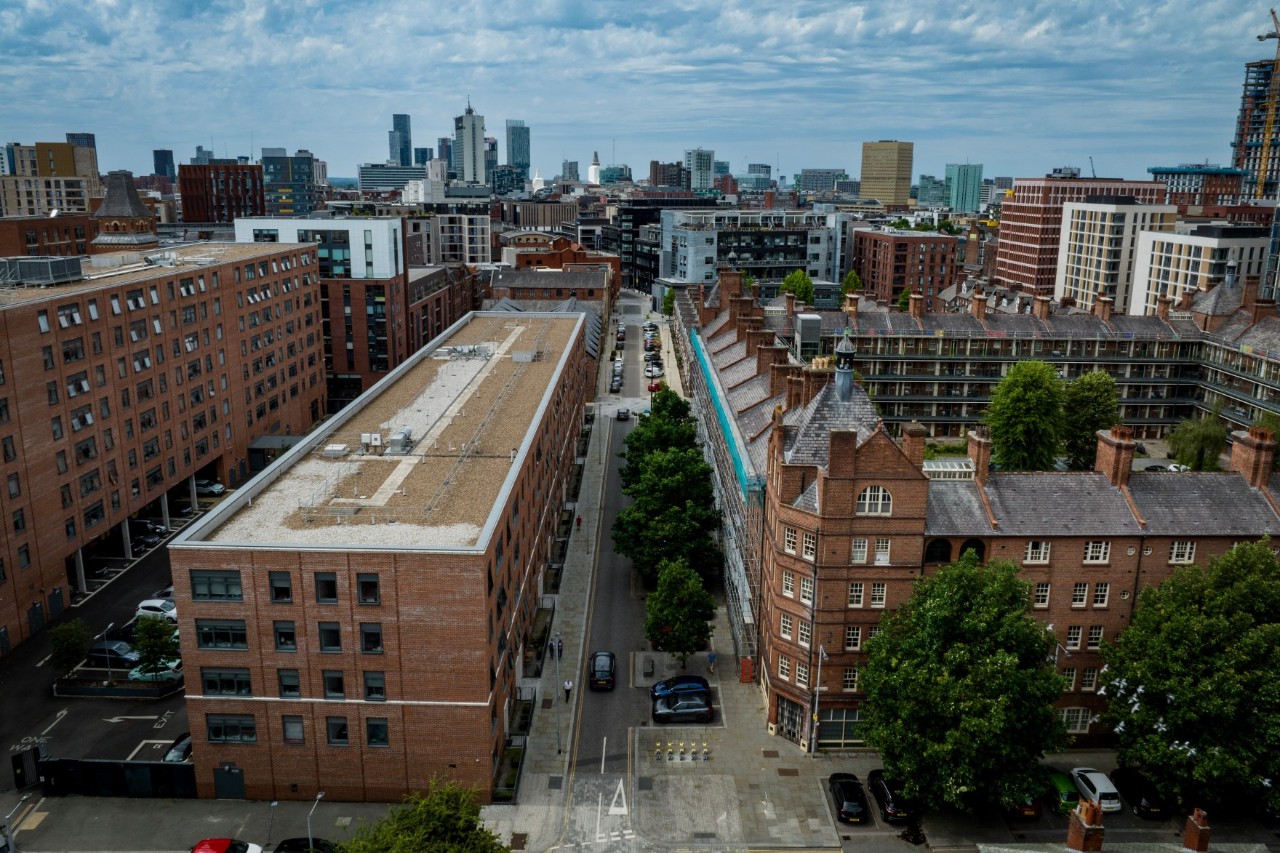 Straßenzug mit rötlich-braunen Backsteingebäuden und Bäumen, im Hintergrund Skyline von Manchester 