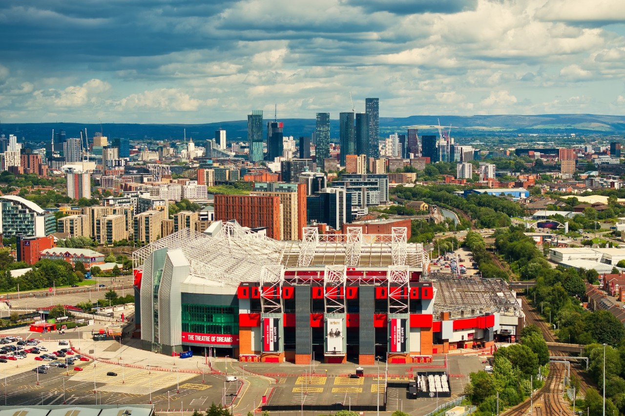 Old Trafford Stadion vor Manchesters Skyline. Bäume säumen die Straßen.