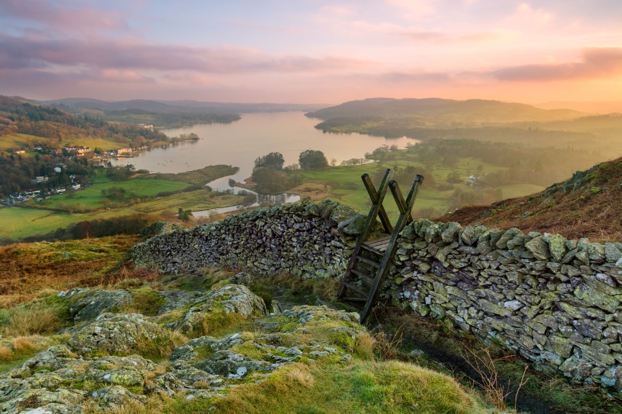 Sonnenaufgang im Lake District, Wiese und Steinmauer mit Holzleiter im Vordergrund, im Hintergrund großer See mit Fluss, hügelige Landschaft, Nebel 