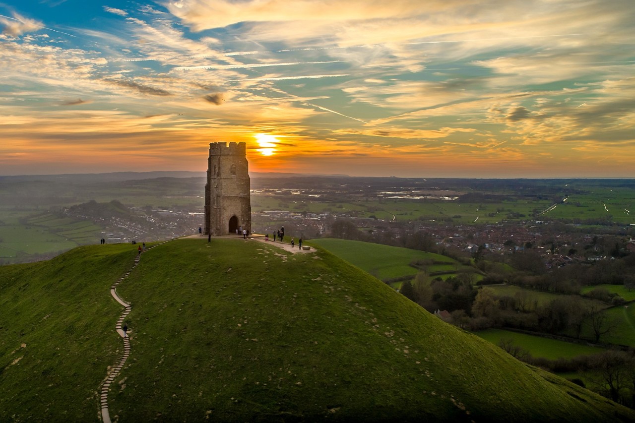 Großer grüner Hügel mit einer historischen Turmruine, umgeben von einer grünen Landschaft. Im Hintergrund ist ein Sonnenuntergang mit beeindruckenden Wolkenformationen zu sehen.