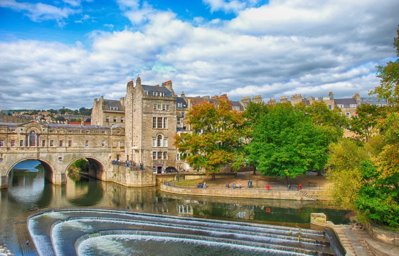 historische Brücke, die über einen Fluss führt. Die Brücke ist von alten Steinhäusern, Bäumen und einer Uferpromenade umgeben. Im Fluss ist ein stufenförmiger Wasserfall integriert.