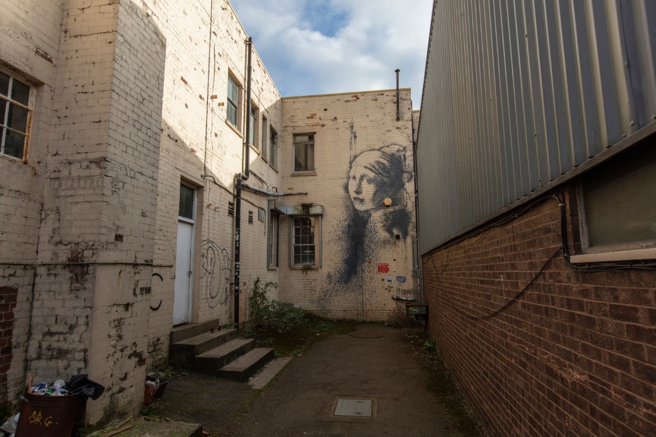 Hinterhof mit hellen Backsteinwänden, die mit Graffiti und Wandkunst verziert sind, darunter ein Porträt eines Mädchens. Rechts ist eine braune Backsteinwand zu sehen, links eine Tür mit vier angrenzenden Stufen.