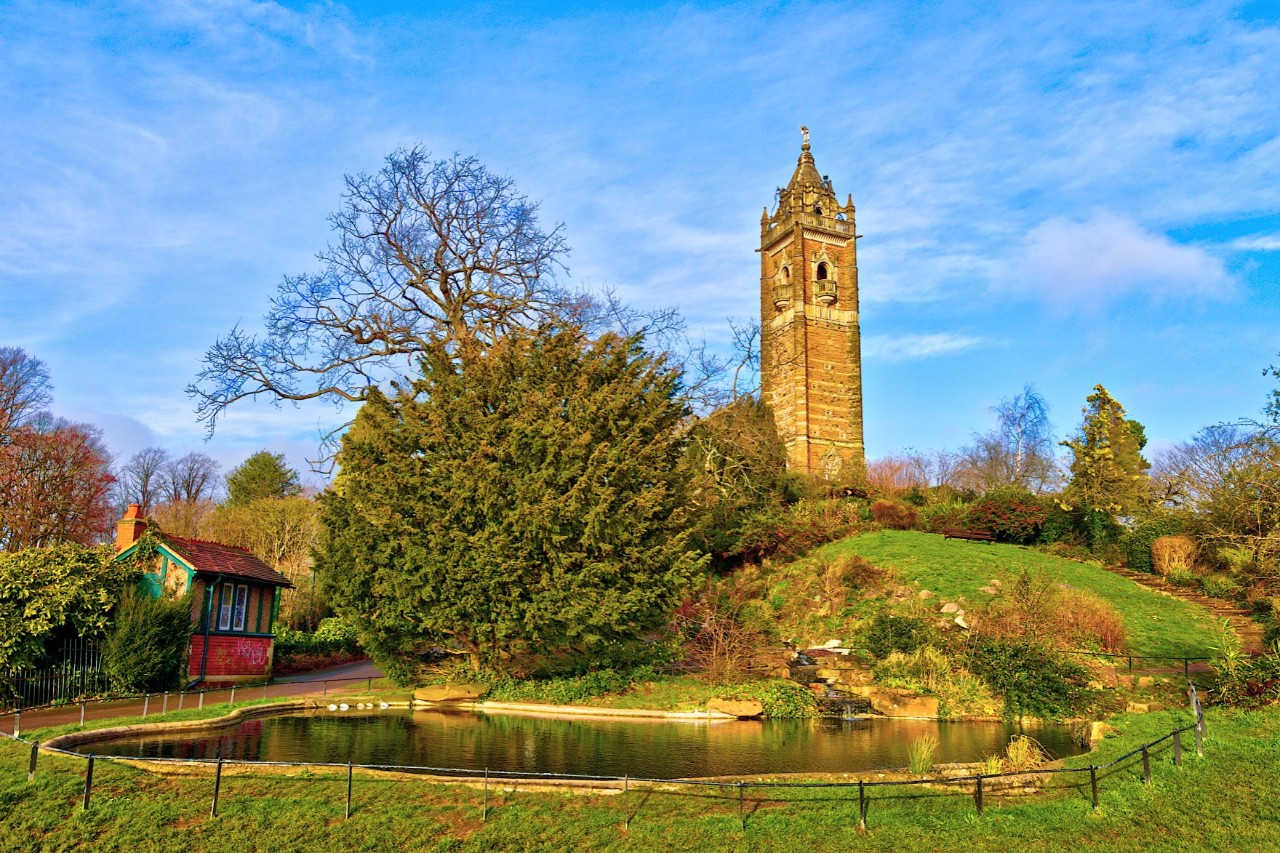Turm auf einem grünen Hügel, umgeben von Bäumen, gepflegten Gärten und einem kleinen Teich im Vordergrund. Links neben dem Teich ist ein kleines Haus zu sehen.