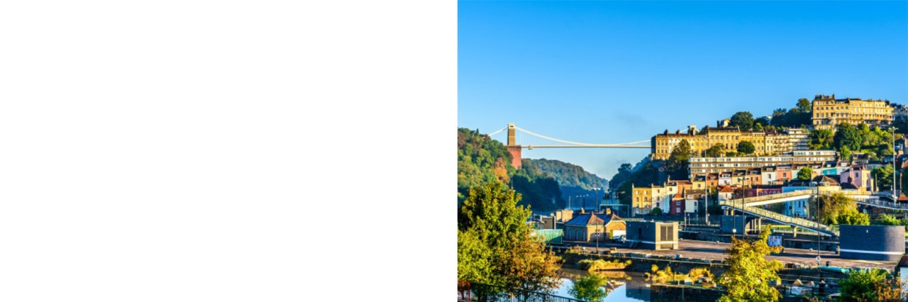 Clifton Suspension Bridge. Im Vordergrund befinden sich Bäume und eine Uferlandschaft, während sich der Stadtteil Clifton mit seinen bunten Häusern auf einem Hügel im Hintergrund erstreckt. 