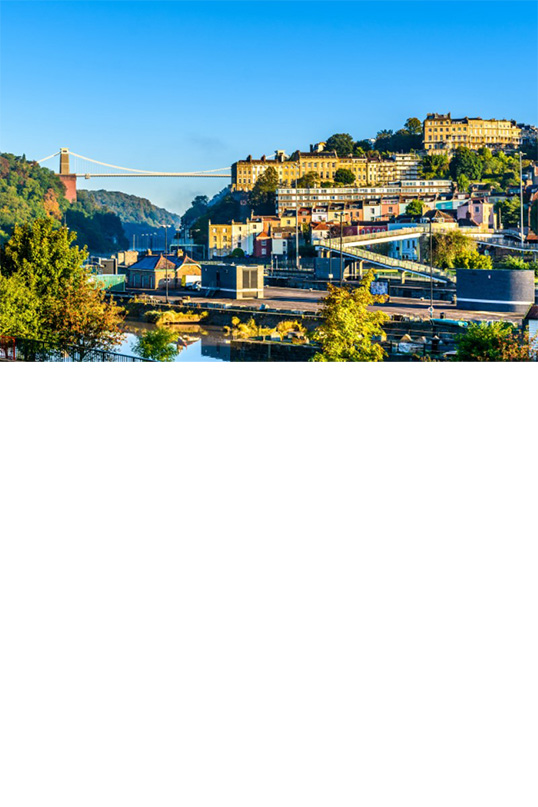 Clifton Suspension Bridge. Im Vordergrund befinden sich Bäume und eine Uferlandschaft, während sich der Stadtteil Clifton mit seinen bunten Häusern auf einem Hügel im Hintergrund erstreckt. 