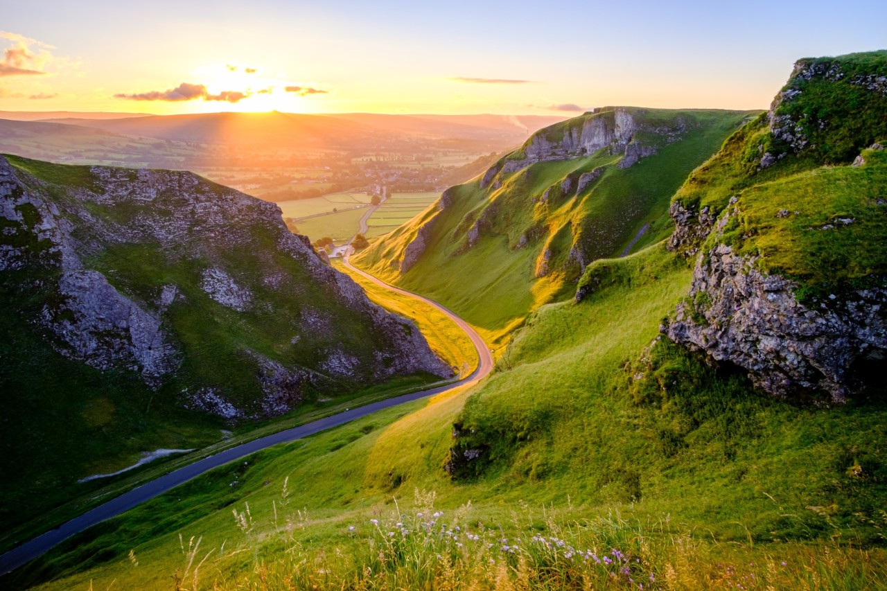 Eine grüne Landschaft bei Sonnenuntergang. Eine kurvige Straße schlängelt sich durch grüne Hügel, die von steilen Felsen gesäumt sind. Im Hintergrund ist ein weites Tal mit Feldern zu sehen. 