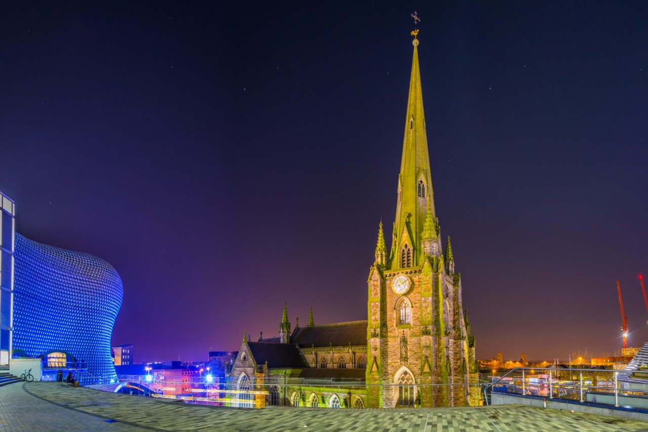 Beleuchtete Kirche mit einem spitzen Turm und einer Uhr in der Nacht. Links davon befindet sich ein modernes, blau beleuchtetes Gebäude mit einer geschwungenen Fassade.