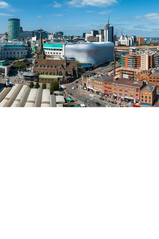 Luftaufnahme von Birmingham: Das Panoramabild zeigt eine dicht bebaute Stadt mit modernen und historischen Gebäuden. Im Vordergrund ist eine belebte Straße und eine Kirche mit spitzem Turm zu sehen. Dahinter ragen Wolkenkratzer und ein großes modernes silberfarbenes, wellenförmiges Haus hervor. Die Szene ist hell erleuchtet und vermittelt ein Gefühl von urbaner Aktivität und Entwicklung. 