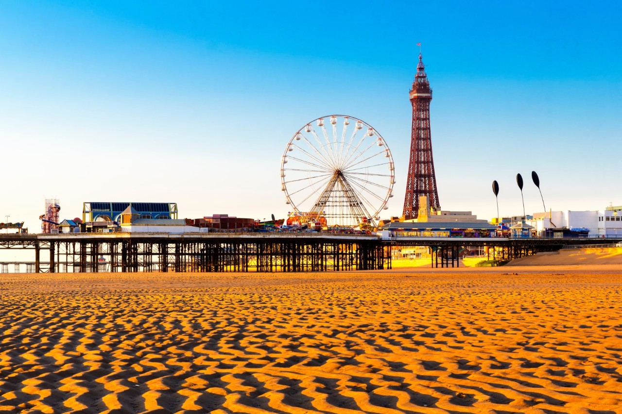 Ein  gepflegter Sandstrand. Dahinter befindet sich eine Seebrücke mit einem Riesenrad und verschiedenen Gebäuden. Rechts steht ein markanter Turm.