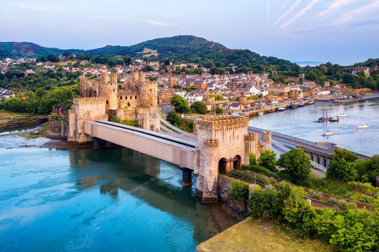 Ene alte Burg, die mit einer Brücke über einen Fluss verbunden ist. Die Burg thront majestätisch über dem Wasser, während im Hintergrund ein Ort mit grünen Hügeln zu sehen ist.