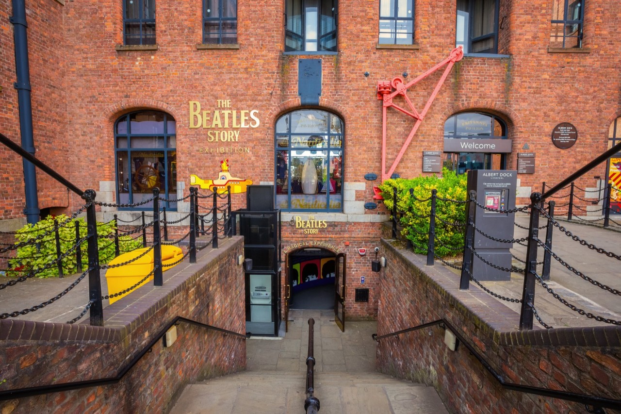 Eingang zur Ausstellung "The Beatles Story". Es handelt sich um ein historisches Backsteingebäude mit Geländern, einer Treppe hinunter zum Eingang und dekorativen Elementen.