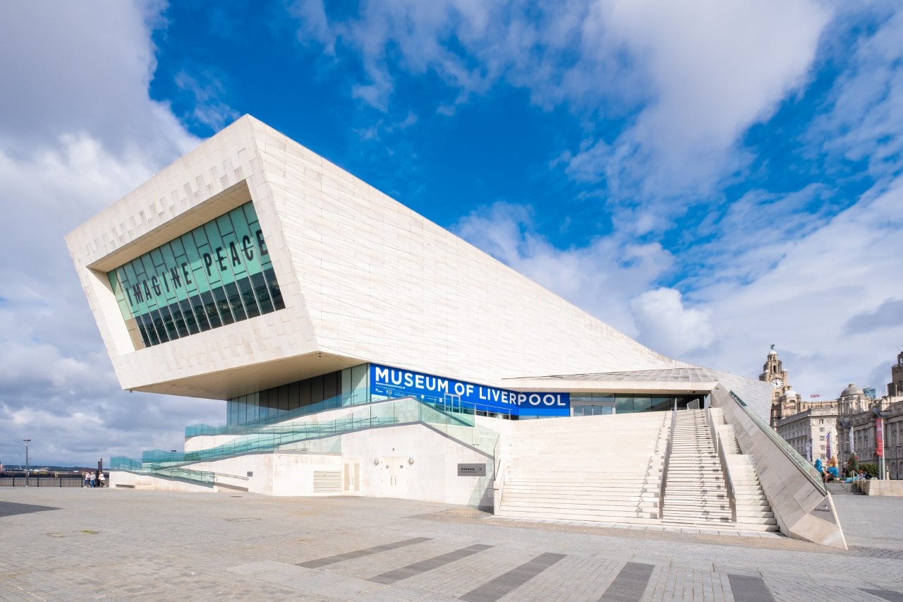 Ein modernes, weißes Gebäude mit geometrischem Design. Über dem Eingang steht "IMAGINE PEACE" in großen Buchstaben. Der blaue Schriftzug weist auf den Namen des Museums hin. 