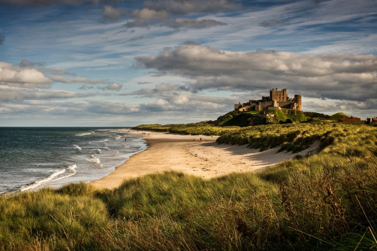 Heller Sandstrand am Meer, der von grün bewachsenen Dünen umrandet ist. Im Hintergrund ist eine Burgkulisse zu sehen.