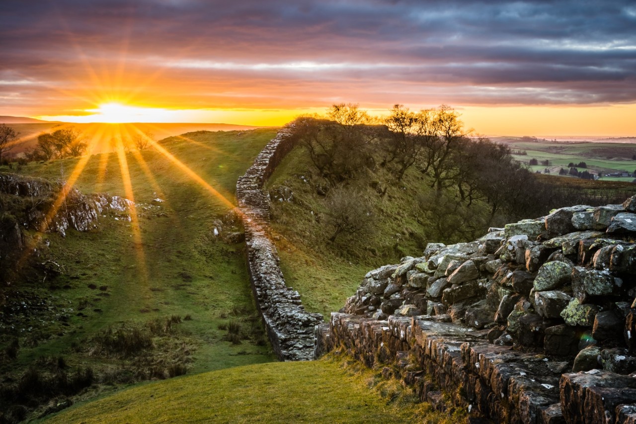 Ein steinerner Wall inmitten einer grünen Hügellandschaft. Die untergehende Sonne taucht das Bild in orange-goldenes Licht.