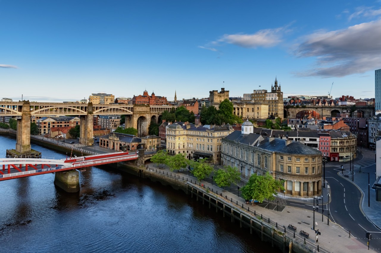 Eine Flussuferpromenade, die von Bäumen gesäumt ist. Dahinter erhebt sich ein Stadtviertel mit zahlreichen Gebäuden. Über den Fluss führt eine rote Brücke auf die andere Seite.