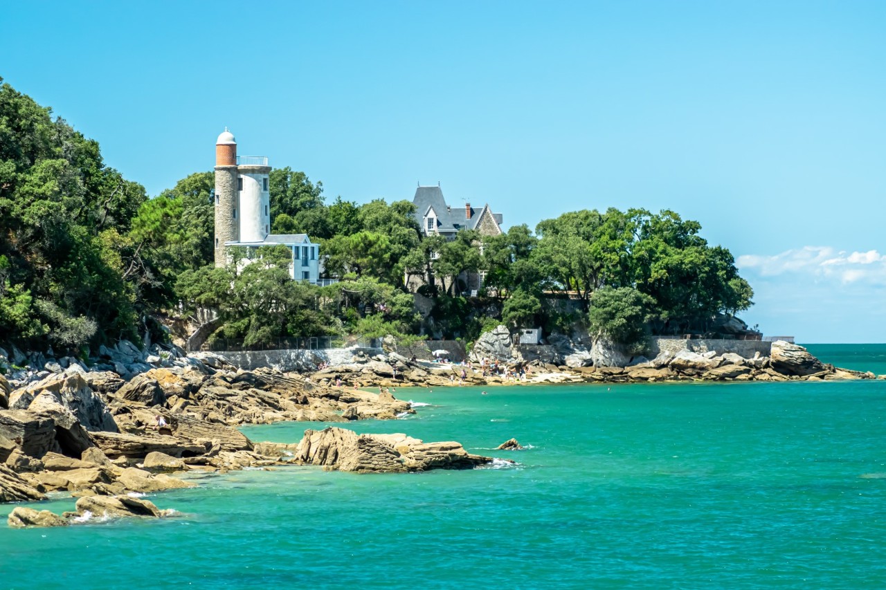 Felsige Küste hinter türkisfarbenem Wasser. Auf den Felsen steht ein Leuchtturm, umgeben von Bäumen und einem historischen Gebäude.