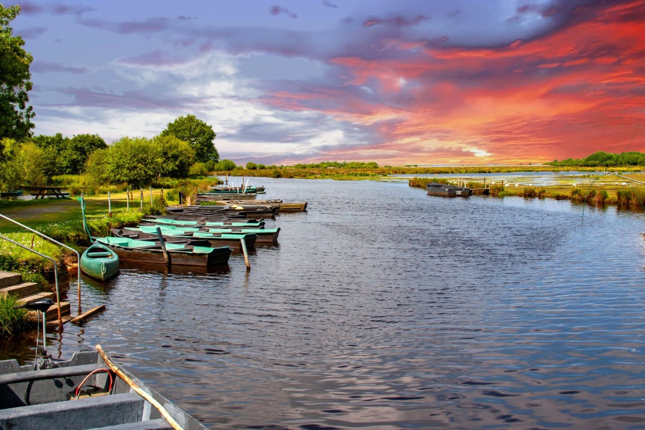 Weitläufige Wasserlandschaft bei Sonnenuntergang zu sehen. Am Ufer des Flusses liegen kleine, grünbraune Boote. Die Umgebung ist von grünen Wiesen und Bäumen geprägt.