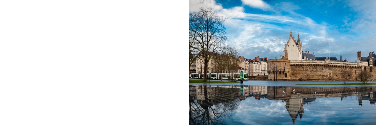 Panoramaaufnahme eines Schlosses, das sich in einem ruhigen Wasserbecken spiegelt und von Häusern umgeben ist. Links ist eine Straßenbahn zu sehen. Die Bäume links und rechts im Bild sind kahl. Der Himmel ist teils von Schleierwolken durchzogen.