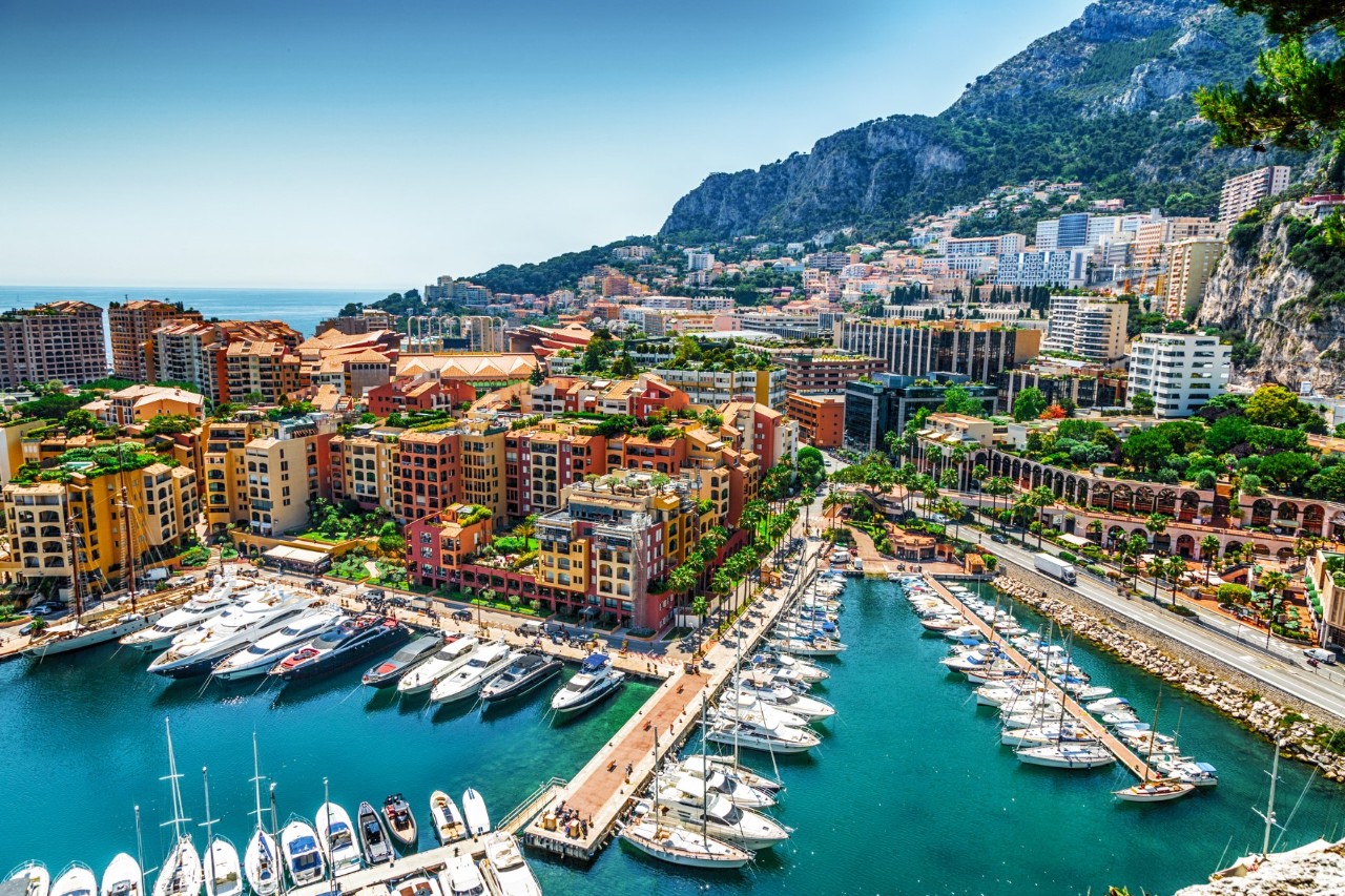 Monte-Carlo mit Yacht-Hafen, bunte Hochhäuser, türkisblaues Wasser, im Hintergrund felsige Berge 