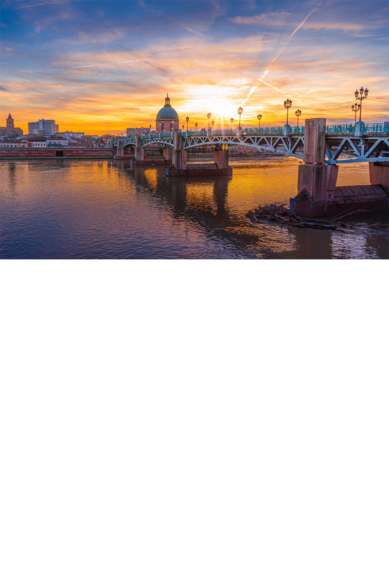 Sonnenuntergang mit Blick auf eine größere Brücke mit Laternen, die über einen Fluss verläuft. Auf der anderen Flussseite sind einige Häuser und die Kuppel einer Kirche zu sehen.