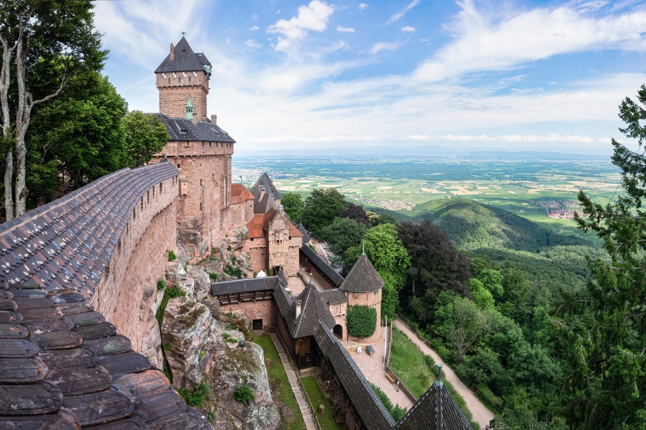 Eine rötlich-sandfarbene Burg mit dunklen Dächern zu sehen, die hoch auf einem Hügel thront. Die Burgmauern und Türme erstrecken sich entlang eines steilen Abhangs. Sie ist von einer grünen Landschaft und bewaldeten Hängen bis zum Horizont umgeben. Der Himmel ist klar und blau.
