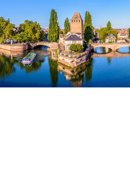 Flusslandschaft mit drei markanten Wehrtürmen, zwei Booten auf dem Fluss, alten Brücken und historischen Gebäuden. Im Hintergrund erhebt sich neben vielen Häusern ein hoher Kirchenturm.