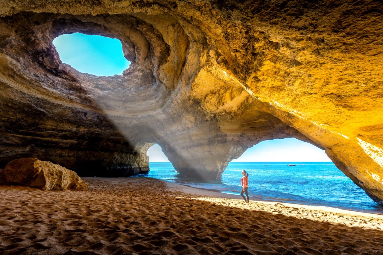 Benagil Höhle mit Felsformationen am Sandstrand des Atlantiks, Licht fällt in die Höhle auf eine Frau, im Hintergrund der Ozean. 