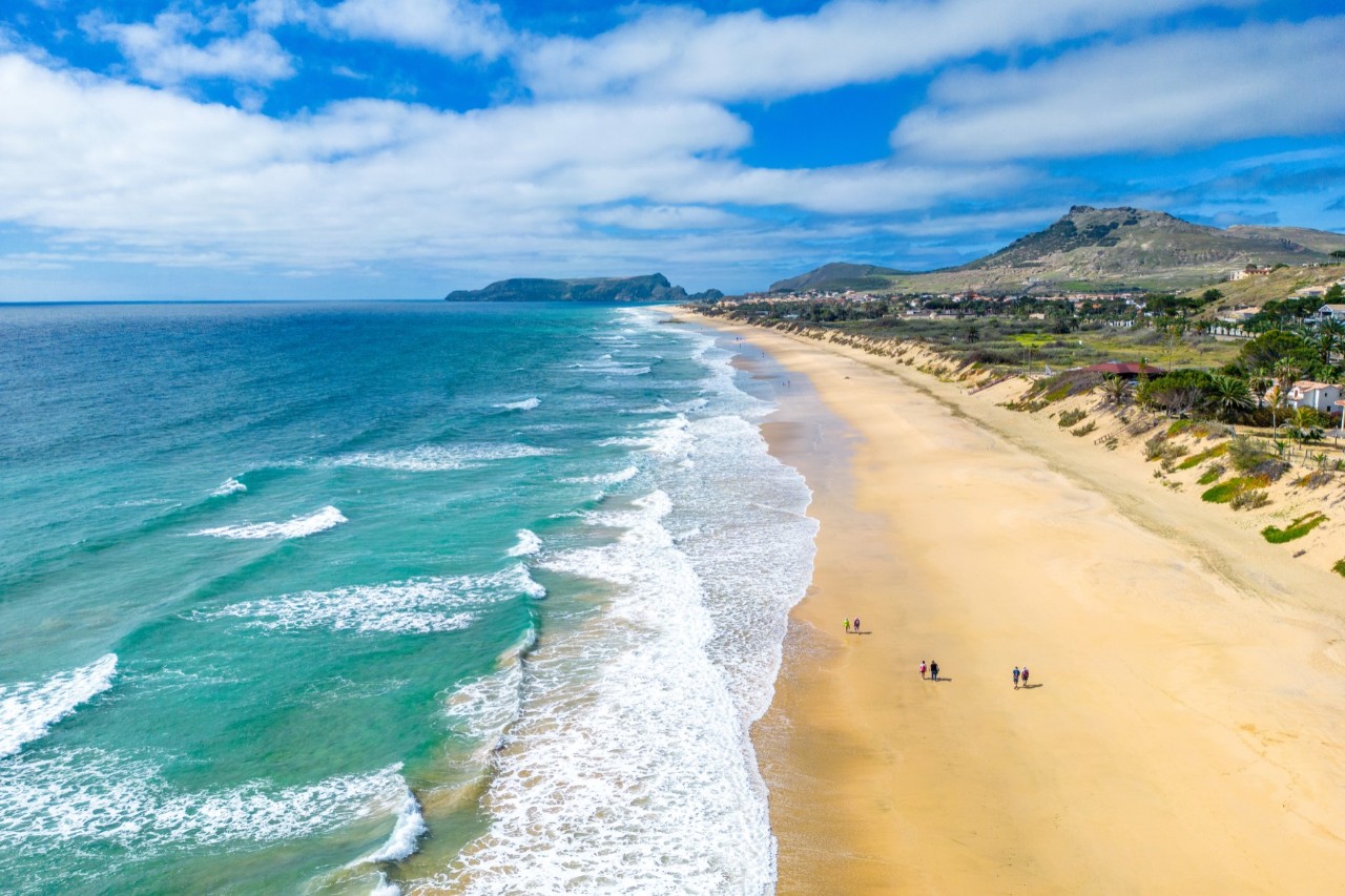 Luftaufnahme eines langen Sandstrandes. Das Meer befindet sich links im Bild. Wellen rollen an den Strand. Links im Bild das grüne Hinterland und Berge 