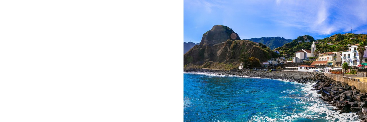 Blaues Meer, im Hintergrund felsige Berge, rechts im Bild eine Uferbefestigung und Dorf mit weißen Häusern. 