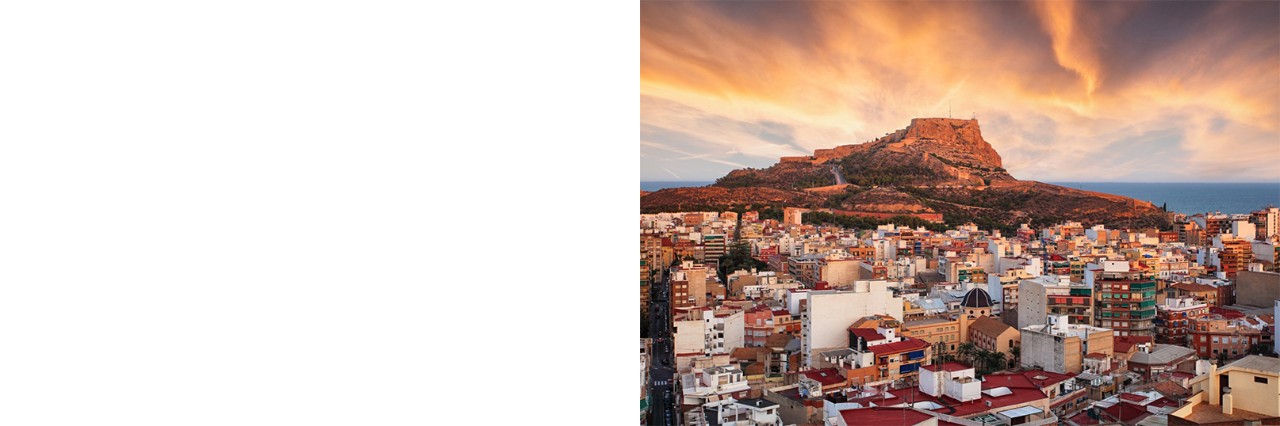 Alicante im Sonnenuntergang. Im Vordergrund ist das vom Sonnenuntergang rötlich erleuchtete Häusermeer zu sehen, im Hintergrund ein Berg mit einer Festung auf dem Gipfel, der ebenfalls rot strahlt.
