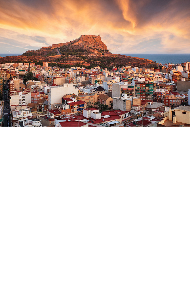 Alicante im Sonnenuntergang. Im Vordergrund ist das vom Sonnenuntergang rötlich erleuchtete Häusermeer zu sehen, im Hintergrund ein Berg mit einer Festung auf dem Gipfel, der ebenfalls rot strahlt.