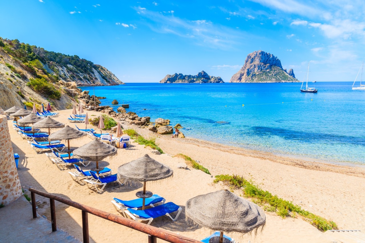 Ein malerischer Strand mit Sonnenschirmen und Liegen, umgeben von Felsen und grünen Hügeln. Im Hintergrund ragen markante Felseninseln aus dem türkisblauen Meer.