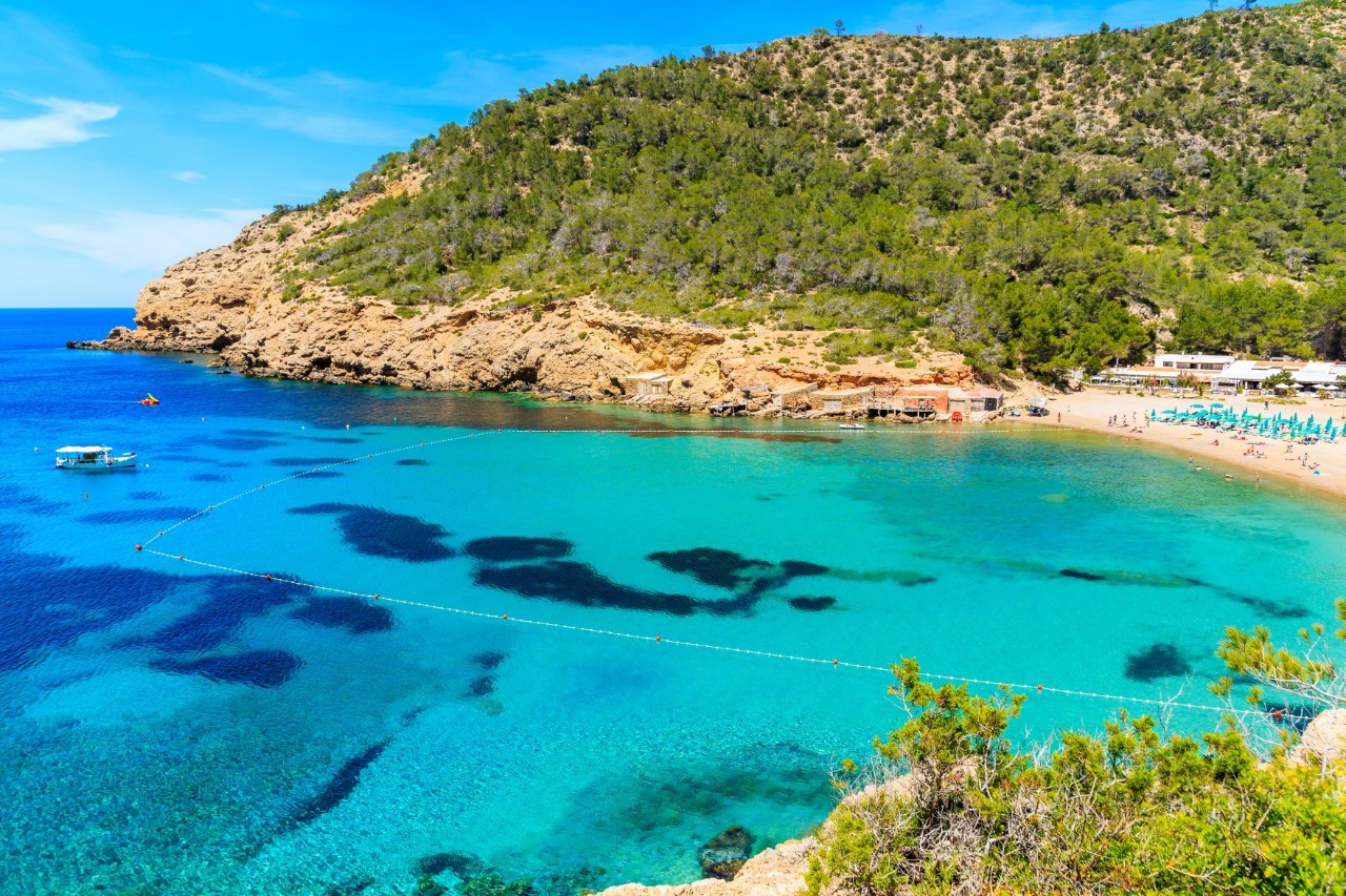 Idyllische Bucht mit klarem, türkisfarbenem Wasser. Am rechten Ufer befindet sich ein Sandstrand mit Sonnenschirmen, während im Hintergrund grüne Hügel und Felsen die Landschaft prägen. 