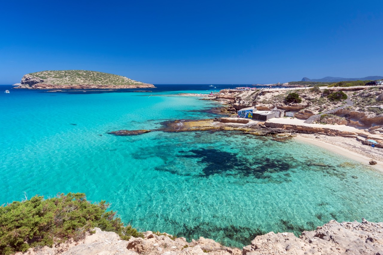 Eine einsame Bucht mit türkisfarbenem, kristallklarem Wasser und einer kleinen Insel im Hintergrund. Der Strand ist von Felsen und karger Vegetation umgeben. 