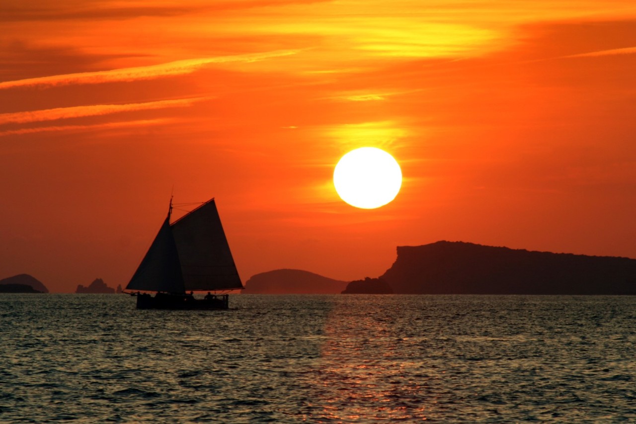 Ein Segelboot bei Sonnenuntergang auf einem Meer. Der Himmel ist in intensiven Orange- und Rottönen gehalten, die Sonne steht tief über dem Horizont. Im Hintergrund sind Inseln zu erkennen.