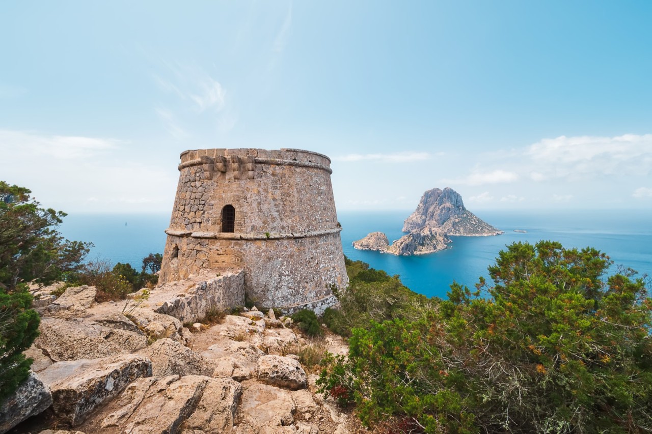 Ein alter sandfarbener Wachturm mit Blick auf einen markanten Felsen, der aus dem Meer ragt. Der Turm ist von grüner Vegetation und sandfarbenen, felsigen Steinen umgeben.