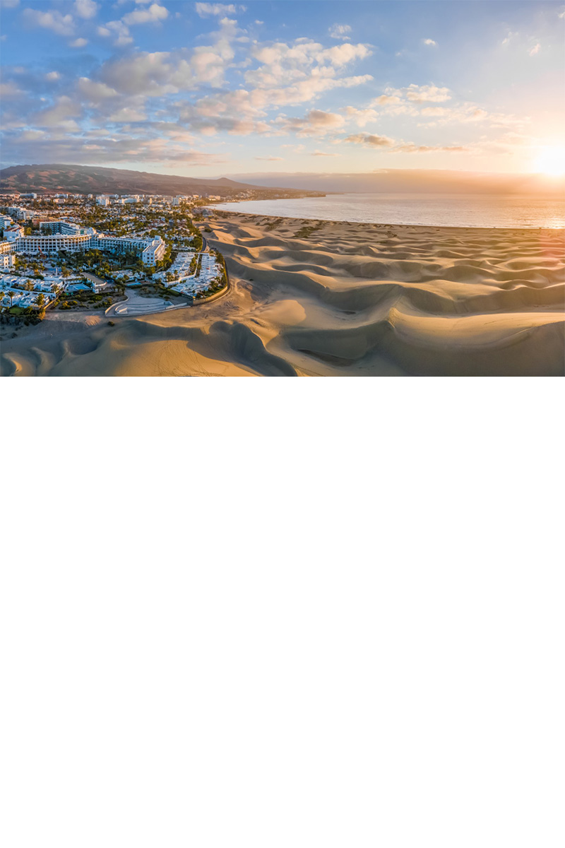 Dünenlandschaft neben Hotel, dahinter Berge und Meer im Sonnenaufgang.