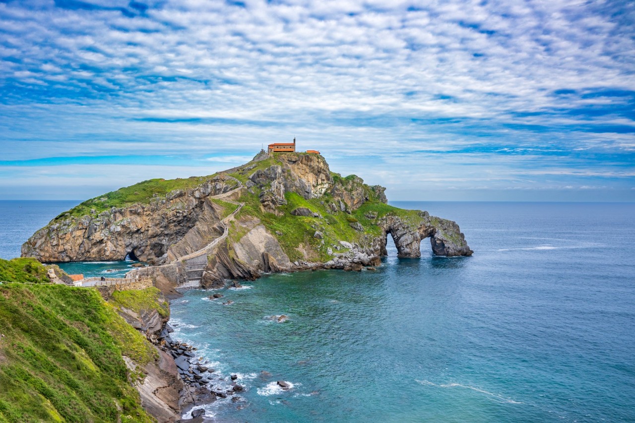 Eine imposante Insel an der Küste, die über eine gewundene Steintreppe erreichbar ist. Auf der Spitze der begrünten Insel steht eine Kapelle. 